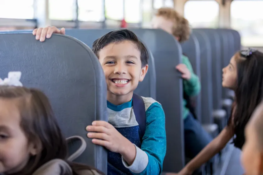 A smiling child looking back over the seat of a school bus, other children in the rows around them