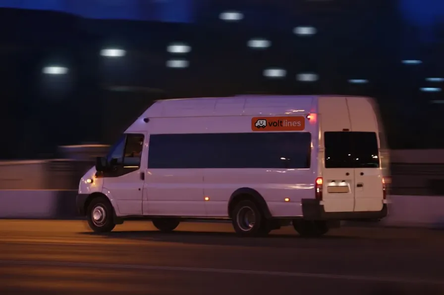 A white Volt Lines minibus running through the city after dark, the lights behind it streaked with motion